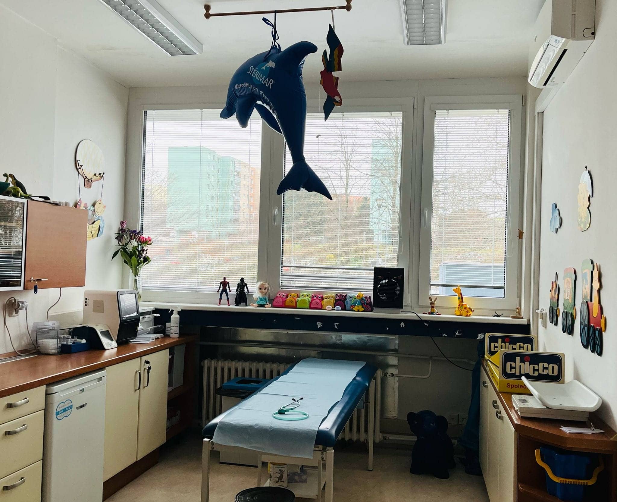 Interiér dětské ordinace s vyšetřovacím lůžkem, pracovním stolem a dekoracemi.