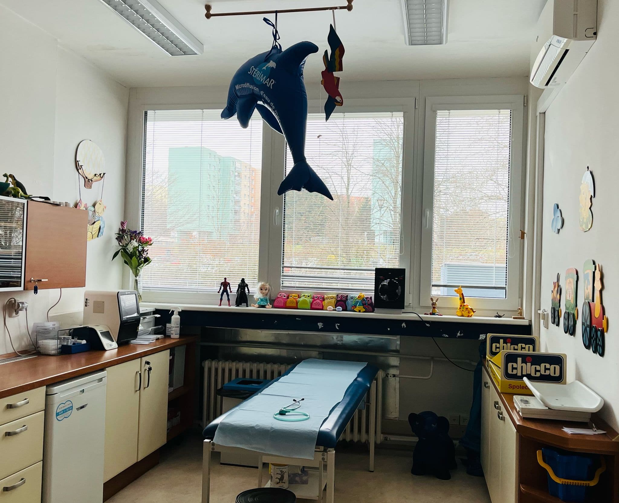 Interiér dětské ordinace s vyšetřovacím lůžkem, pracovním stolem a dekoracemi.